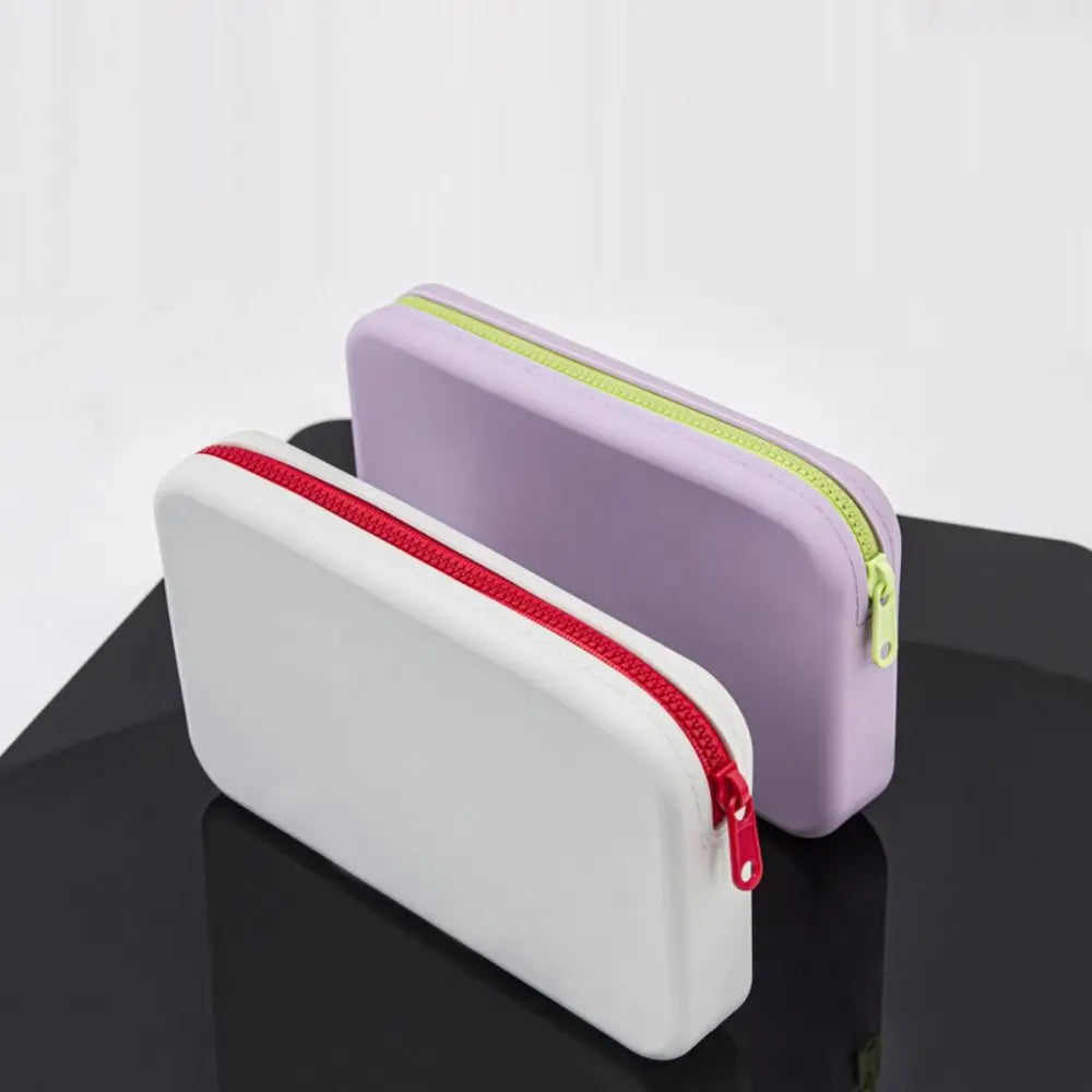 Due astucci rettangolari in silicone, uno bianco con cerniera rossa e uno lilla con cerniera verde, su sfondo nero. Accessori scuola moderni.