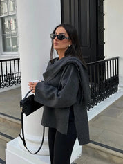 Donna con occhiali da sole indossa cappotto grigio elegante, borsa nera a tracolla, posa su sfondo urbano. Moda autunnale, stile chic.