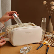 Borsa cosmetica beige con flacone spray trasparente, pennelli trucco e profumo su tavolo marrone. Elegante e pratica per organizzare prodotti di bellezza.