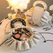 Borse cosmetiche beige con manici, contenenti pennelli trucco e palette ombretti, su tavolo con lampada piumata e fiori. Accessori bellezza eleganti.
