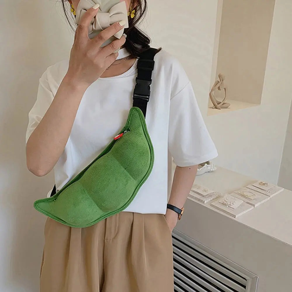 Donna con borsa a tracolla verde a forma di baccello di pisello, indossa maglietta bianca e pantaloni beige. Accessorio moda originale e trendy.