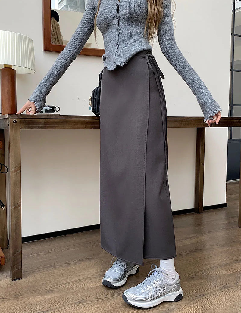 Donna con maglione grigio e gonna lunga marrone, scarpe da ginnastica argentate, in un ambiente interno moderno con tavolo in legno e lampada.