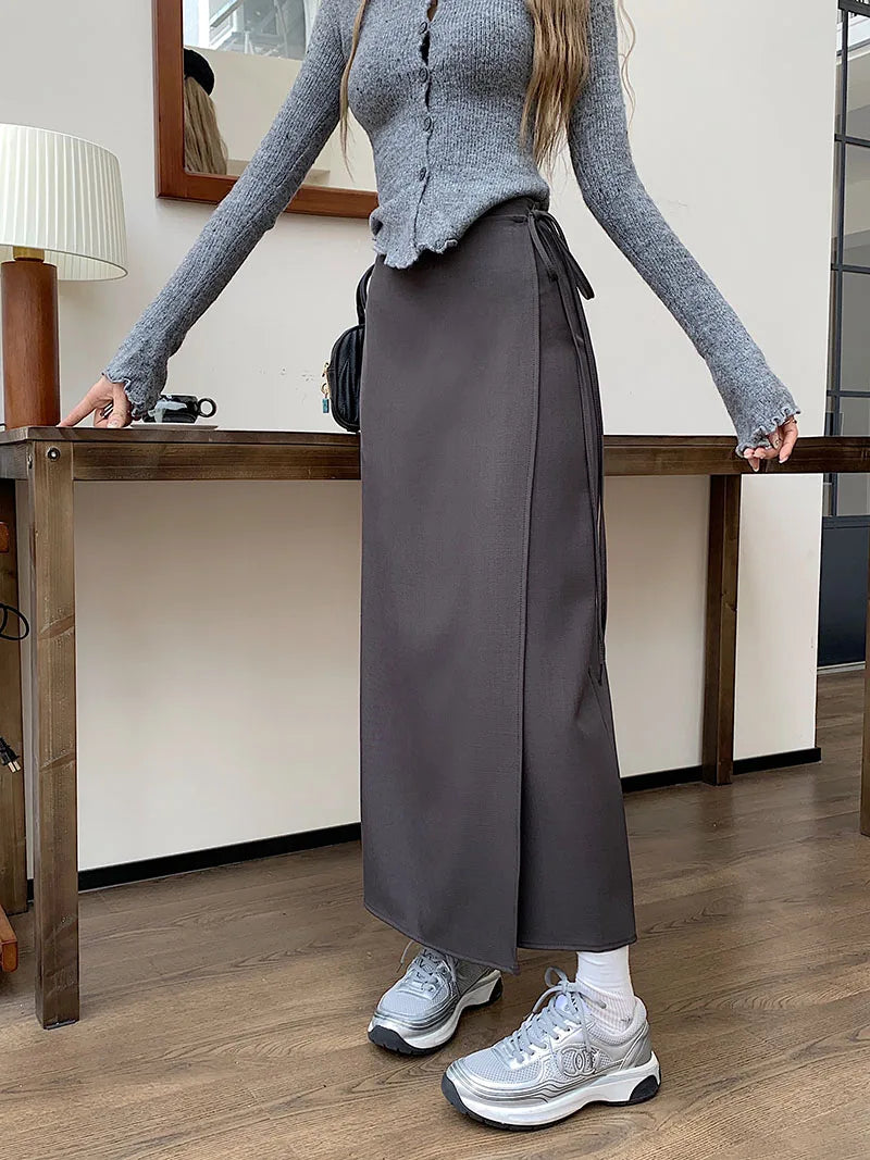 Donna con maglione grigio e gonna lunga marrone, scarpe da ginnastica argentate, in un ambiente interno moderno con tavolo in legno e lampada.