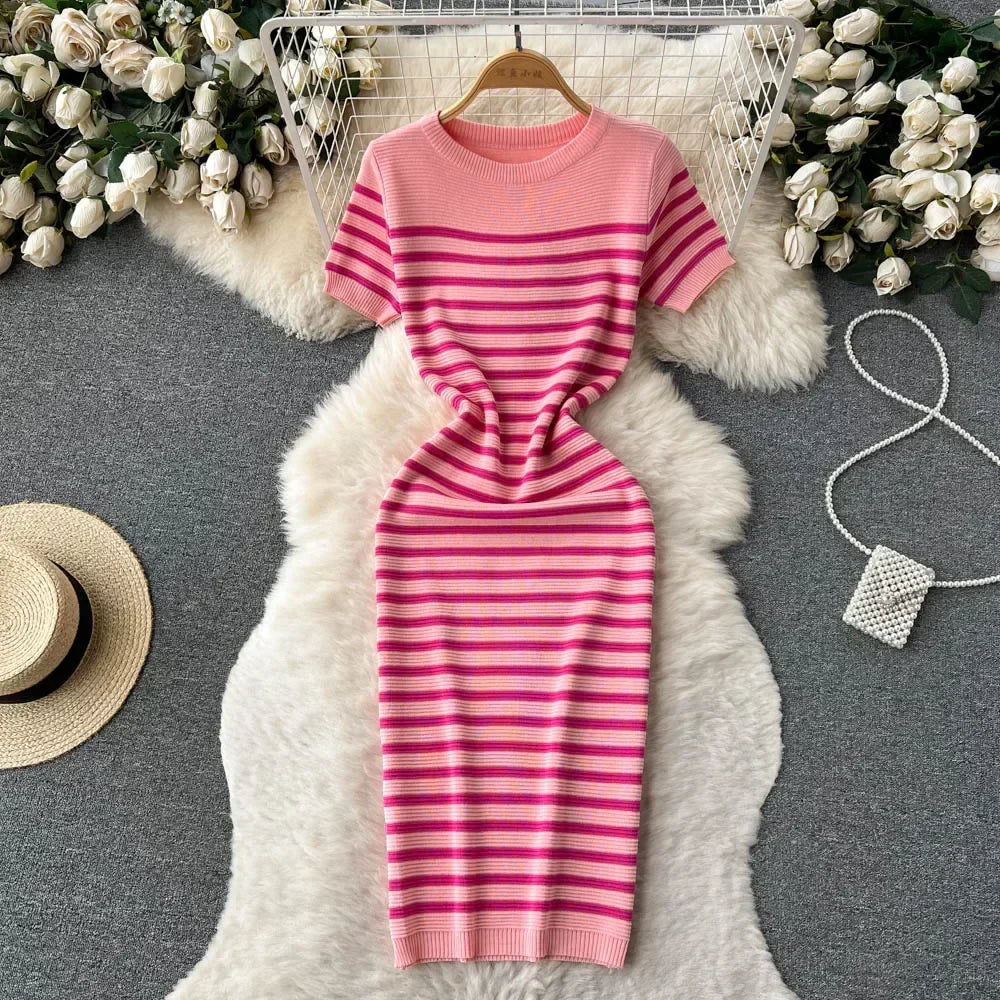 Abito a righe rosa e fucsia su tappeto bianco, circondato da rose bianche e accessori moda. Elegante e moderno, perfetto per la primavera.