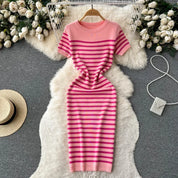 Abito a righe rosa e fucsia su tappeto bianco, circondato da rose bianche e accessori moda. Elegante e moderno, perfetto per la primavera.