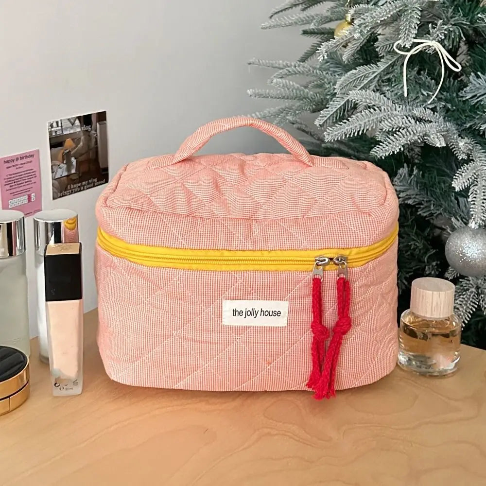 Beauty case rosa a righe con cerniera gialla e manico, su tavolo accanto a profumi e albero di Natale. Accessorio da viaggio elegante e pratico.