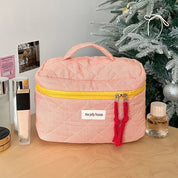Beauty case rosa a righe con cerniera gialla e manico, su tavolo accanto a profumi e albero di Natale. Accessorio da viaggio elegante e pratico.