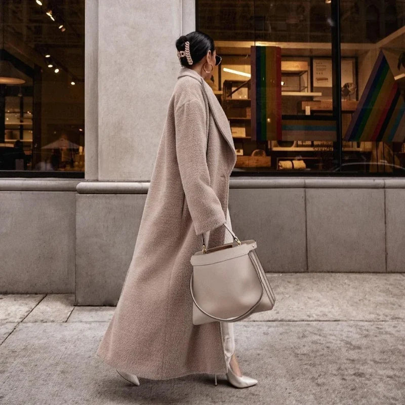 Donna con cappotto lungo beige, borsa elegante e tacchi alti cammina in città. Moda autunnale, stile urbano, abbigliamento chic.