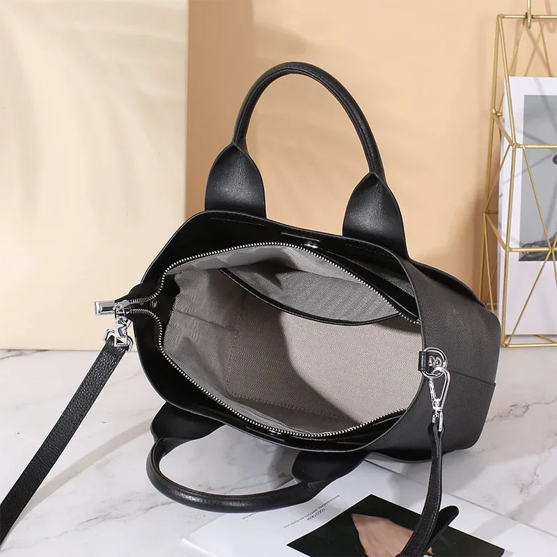 Borsa a mano nera in pelle con interno beige, cerniera aperta, manici robusti e tracolla regolabile, su sfondo chiaro. Accessorio moda elegante.