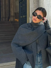Donna con occhiali da sole neri, cappotto grigio e jeans, posa davanti a un edificio storico. Moda autunnale elegante, stile urbano chic.