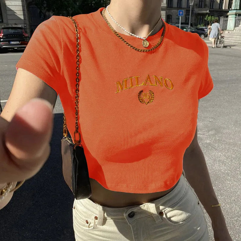 Donna con t-shirt arancione "Milano", collana dorata, borsa a tracolla, jeans chiari. Moda estiva urbana, abbigliamento casual trendy.