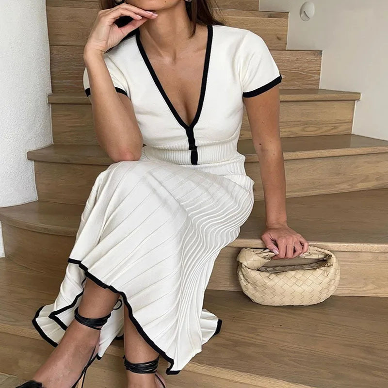 Donna seduta su scala in abito bianco a coste con scollo a V e dettagli neri, borsa intrecciata beige, sandali neri con tacco. Moda elegante casual.
