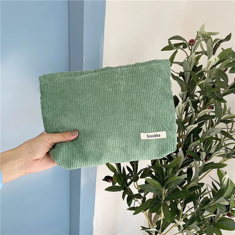 Cuscino verde in velluto a coste, tenuto in mano, accanto a una pianta decorativa. Design moderno, ideale per arredamento casa.