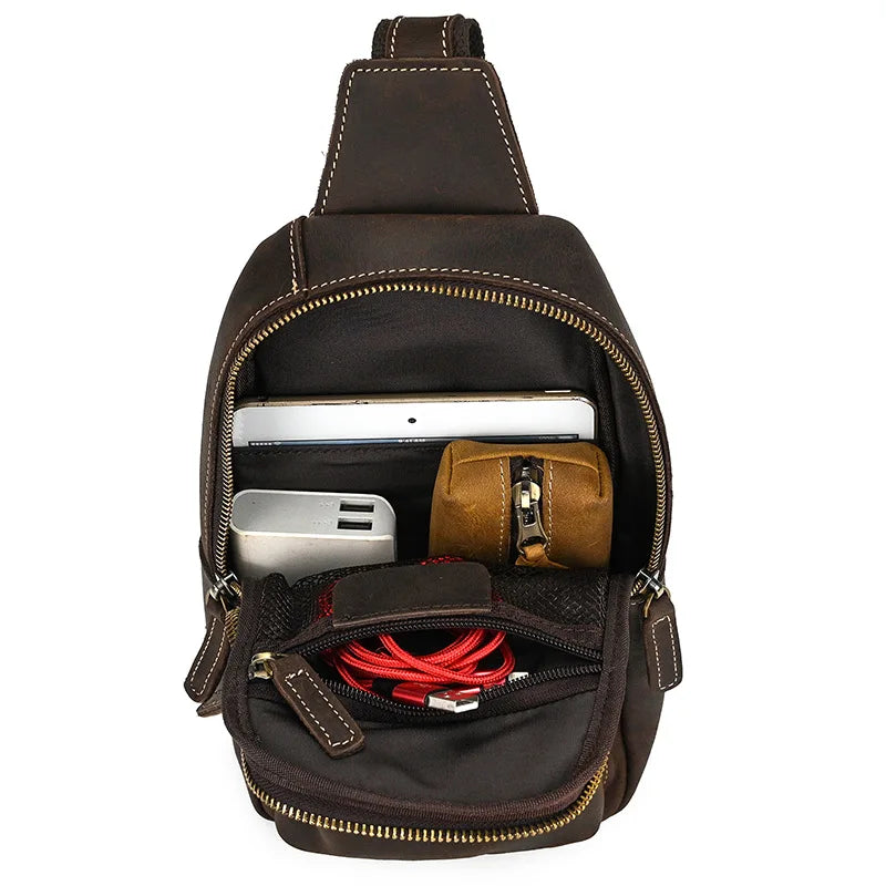 Zaino a tracolla in pelle marrone con tasche aperte, contenente tablet, power bank, cavi e accessori. Design compatto e funzionale per viaggi e lavoro.