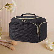 Borsa cosmetica nera trapuntata con cerniere dorate, manico superiore, fiori bianchi e pennelli trucco su sfondo beige. Elegante e funzionale.