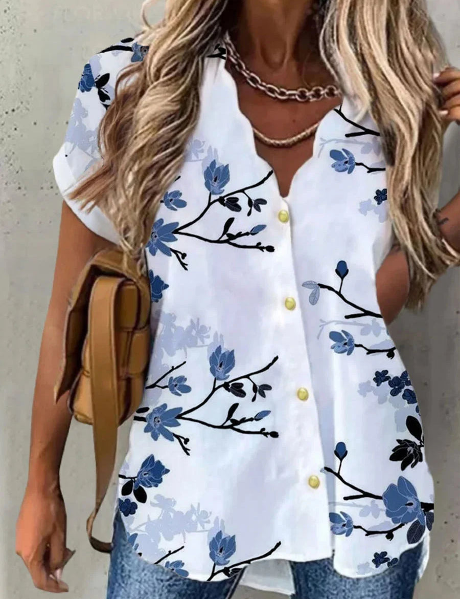 Camicia donna bianca con stampa floreale blu, maniche corte, bottoni gialli, abbinata a jeans e borsa marrone. Moda estiva casual elegante.