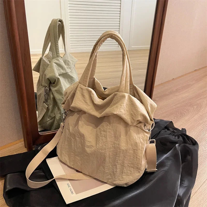 Borsa a spalla beige in tessuto morbido, design casual, con manici e tracolla regolabile, riflessa in uno specchio su sfondo di legno chiaro.