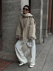 Donna con cappotto beige, pantaloni bianchi e borsa intrecciata, appoggiata a un muro di pietra. Look elegante e moderno, moda autunnale.