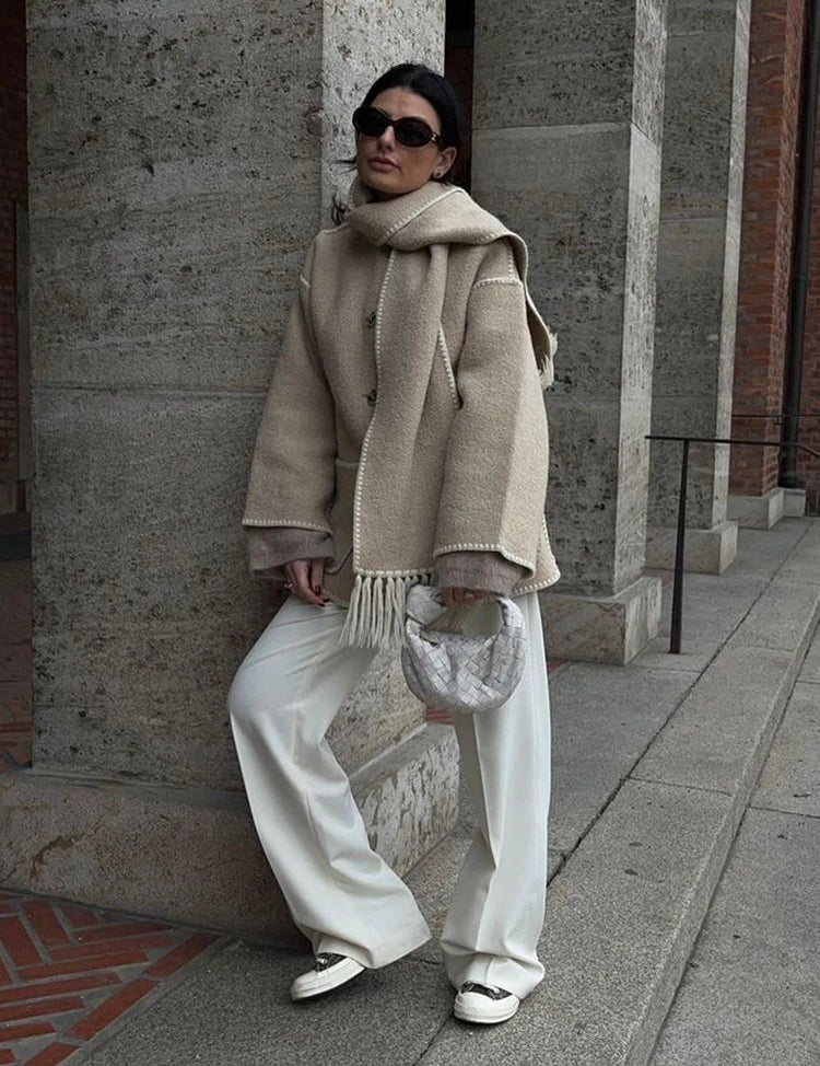 Donna con cappotto beige, pantaloni bianchi e borsa intrecciata, appoggiata a un muro di pietra. Look elegante e moderno, moda autunnale.