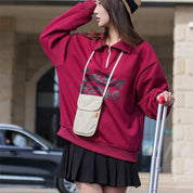 Donna con felpa rossa oversize, gonna nera e cappello di paglia, tiene una valigia. Moda casual autunnale, street style femminile.