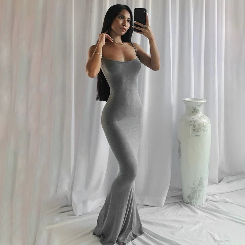 Donna in abito lungo grigio aderente, posa davanti a tende bianche con vaso decorativo. Moda elegante, abbigliamento femminile, stile sofisticato.