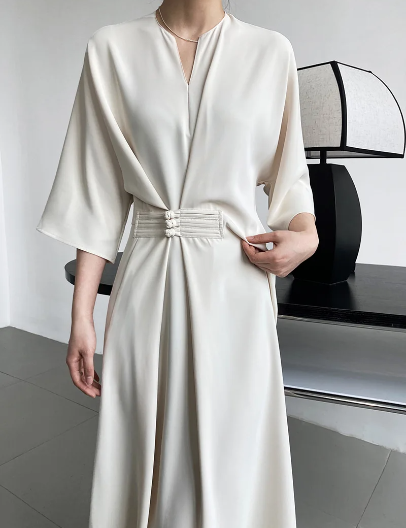 Abito elegante bianco avorio con cintura decorativa, maniche ampie e scollo a V, indossato da una donna. Design minimalista e moderno.