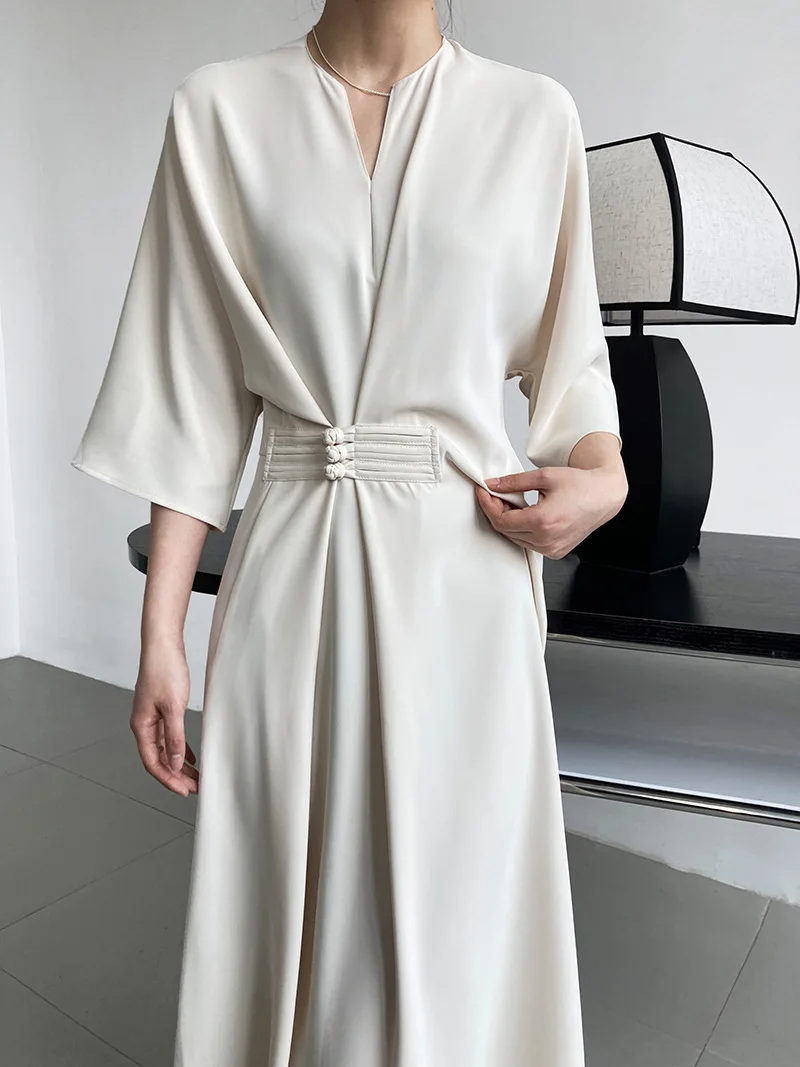 Abito elegante bianco avorio con cintura decorativa, maniche ampie e scollo a V, indossato da una donna. Design minimalista e moderno.