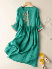 Abito verde in lino con ricami floreali, maniche a tre quarti, abbinato a borsa trasparente e sandali gialli. Elegante e casual.