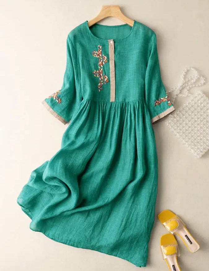 Abito verde in lino con ricami floreali, maniche a tre quarti, abbinato a borsa trasparente e sandali gialli. Elegante e casual.