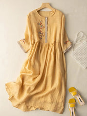 Abito lungo in lino giallo con ricami floreali, maniche a tre quarti, scollo rotondo. Accessori: borsa trasparente, sandali gialli.