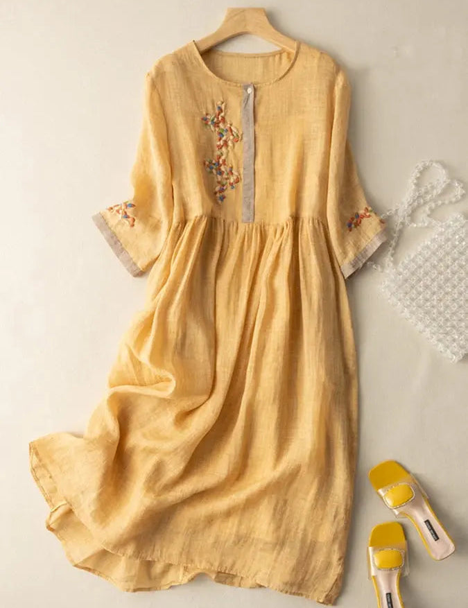 Abito lungo in lino giallo con ricami floreali, maniche a tre quarti, scollo rotondo. Accessori: borsa trasparente, sandali gialli.