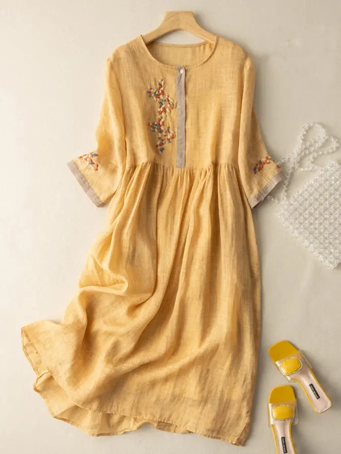 Abito lungo in lino giallo con ricami floreali, maniche a tre quarti, scollo rotondo. Accessori: borsa trasparente, sandali gialli.