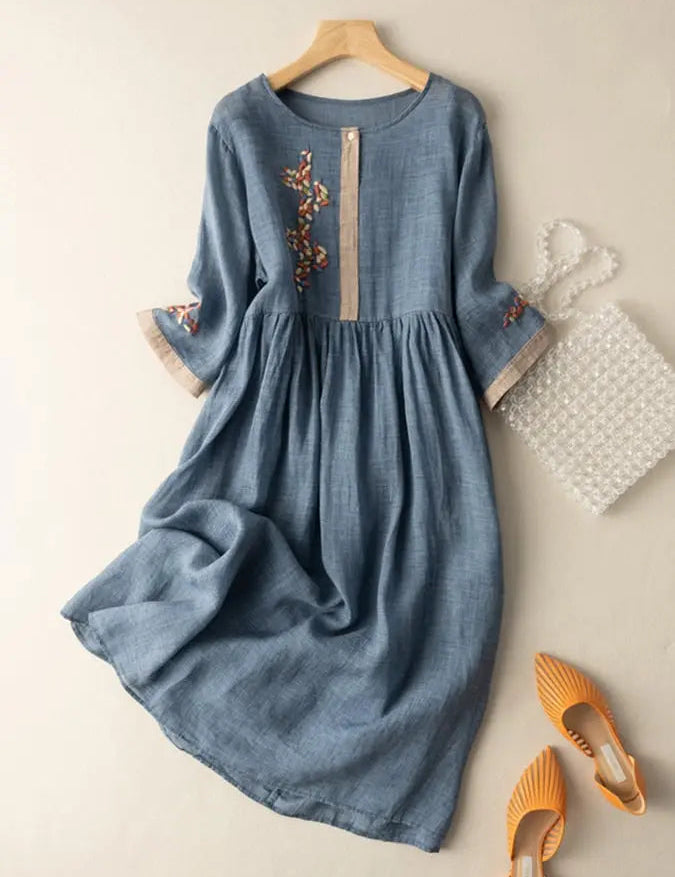 Abito in lino blu con ricami floreali, maniche a tre quarti, abbinato a borsa trasparente e scarpe arancioni. Moda donna elegante e casual.