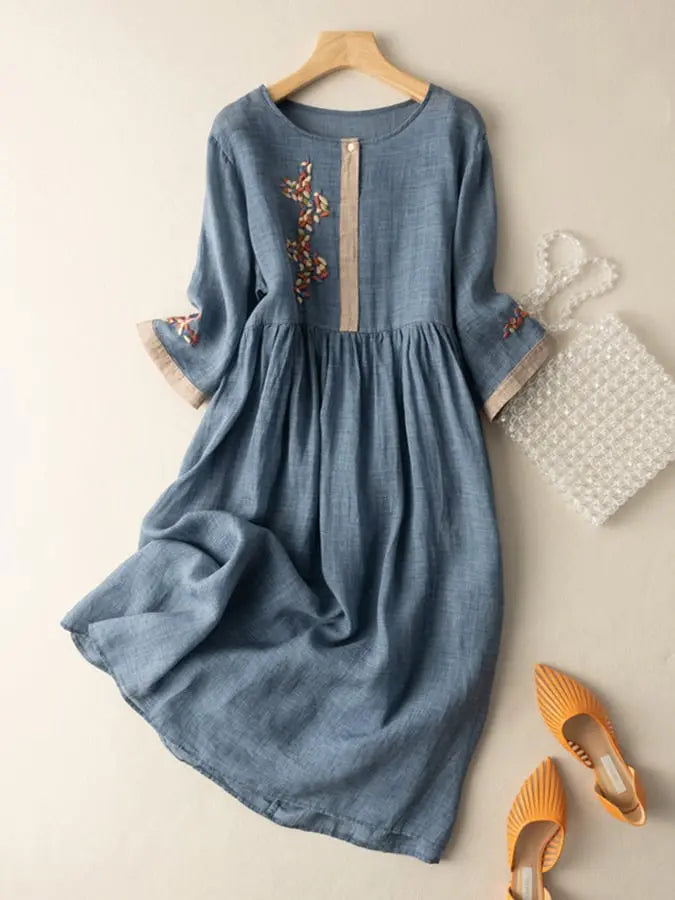 Abito in lino blu con ricami floreali, maniche a tre quarti, abbinato a borsa trasparente e scarpe arancioni. Moda donna elegante e casual.
