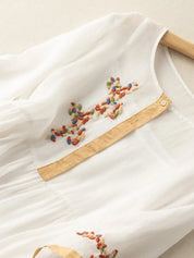 Blusa bianca in chiffon con ricami colorati, dettagli beige, maniche lunghe, design elegante e leggero. Perfetta per look estivi e casual.