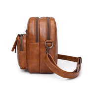 Zaino in pelle marrone con doppia cerniera, tasche laterali e tracolla regolabile. Design elegante e pratico per uso quotidiano.