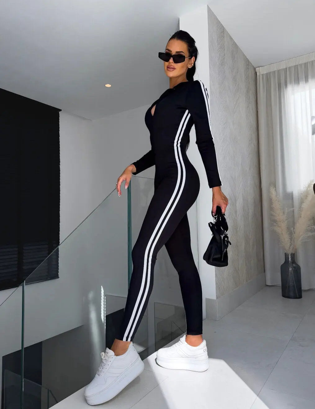 Donna in tuta sportiva nera con strisce bianche, occhiali da sole e scarpe da ginnastica bianche, posa moderna in interni eleganti.