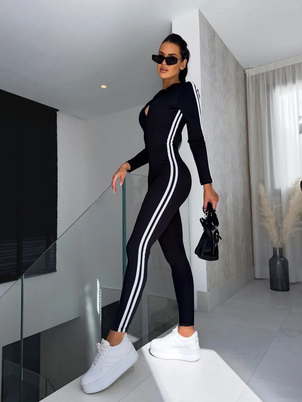 Donna in tuta sportiva nera con strisce bianche, occhiali da sole e scarpe da ginnastica bianche, posa moderna in interni eleganti.