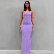 Abito lungo aderente viola, senza maniche, con scollo quadrato, indossato da una donna su sfondo grigio. Elegante e moderno, perfetto per eventi serali.