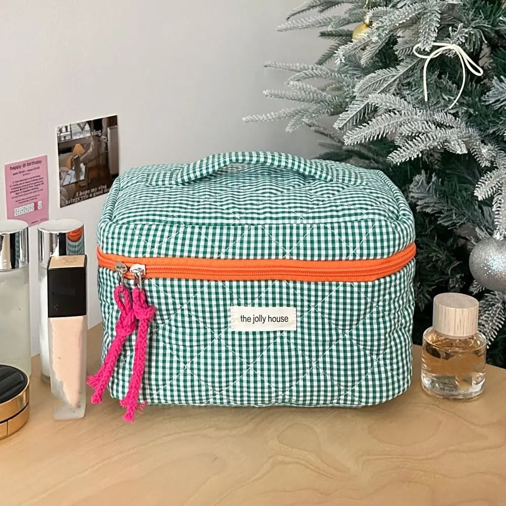 Borsa cosmetica a quadretti verdi con cerniera arancione su tavolo di legno, accanto a prodotti di bellezza e albero di Natale decorato.