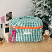 Borsa cosmetica a quadretti verdi con cerniera arancione su tavolo di legno, accanto a prodotti di bellezza e albero di Natale decorato.