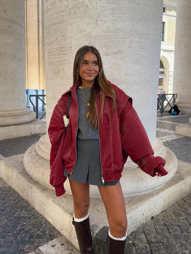 Giovane donna con giacca oversize rossa, maglia grigia e gonna corta, posa davanti a colonne storiche. Moda autunnale casual chic.