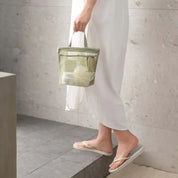 Donna con borsa da doccia trasparente verde, contenente prodotti per l'igiene, su gradino in bagno. Indossa abito bianco e infradito.