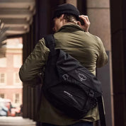 Uomo con zaino a tracolla Osprey nero, giacca verde e berretto nero, cammina in città. Accessorio moda urbano, pratico e resistente.