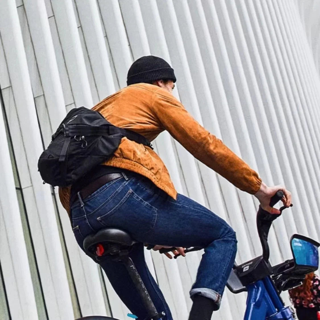 Uomo in bicicletta con giacca marrone, jeans e zaino nero, davanti a edificio moderno. Ciclismo urbano, moda casual, trasporto ecologico.