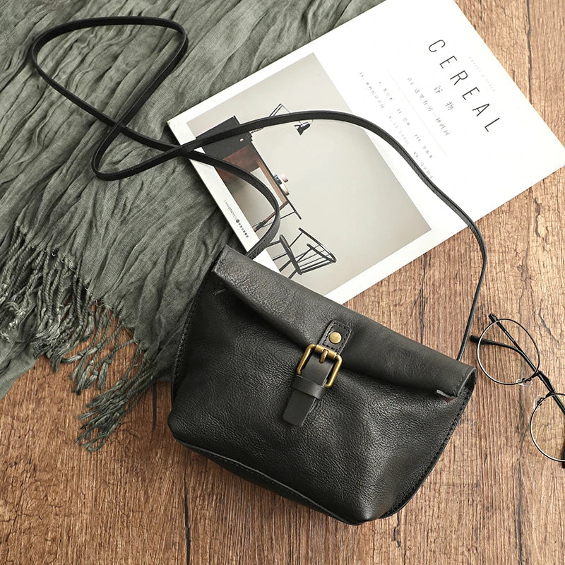 Borsa a tracolla in pelle nera su tavolo di legno, accanto a occhiali e rivista. Design elegante con fibbia dorata. Accessorio moda donna.