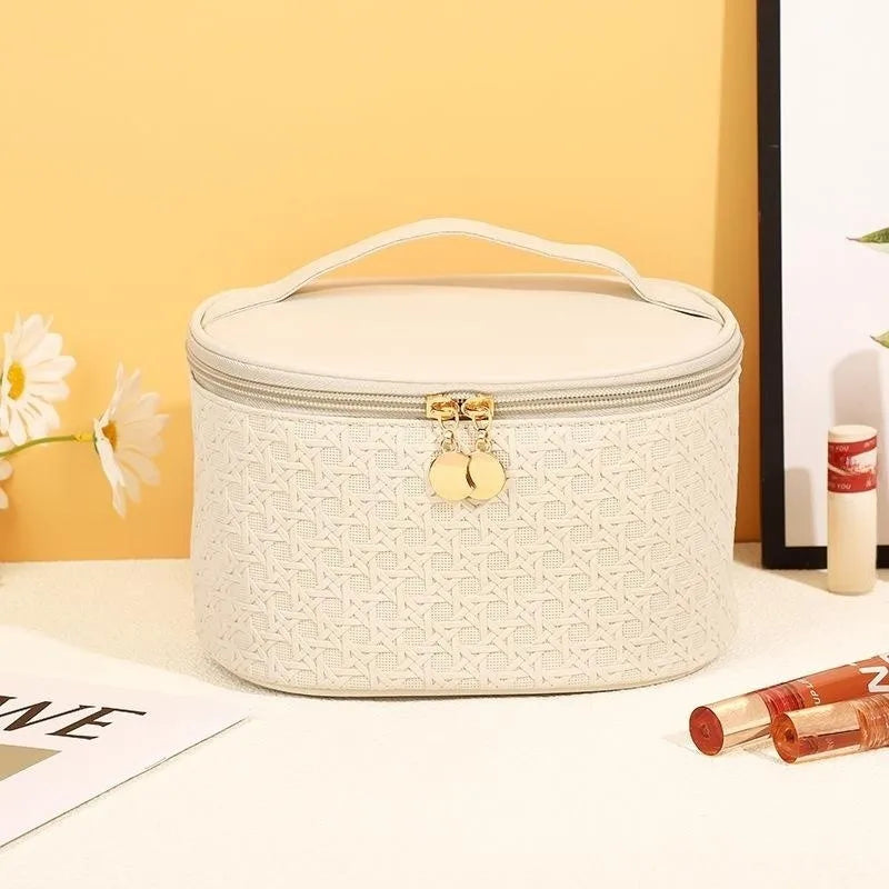 Astuccio cosmetico bianco con motivo intrecciato, chiusura a zip dorata, manico superiore. Accessori trucco e fiori sullo sfondo.