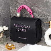 Borsa trucco nera con scritta "Personal Care", manico rosa con perla, su lavandino in marmo. Accessori bagno: dispenser sapone, profumo, fiori.