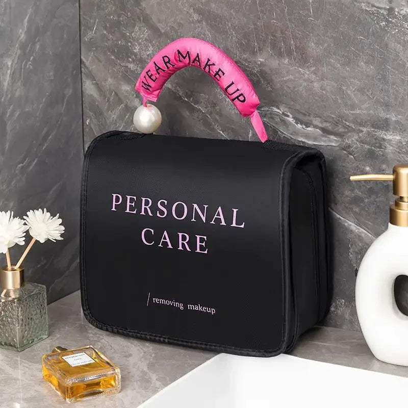 Borsa trucco nera con scritta "Personal Care", manico rosa con perla, su lavandino in marmo. Accessori bagno: dispenser sapone, profumo, fiori.