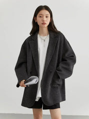 Donna con giacca oversize grigia, maglietta bianca e pantaloncini neri. Moda casual autunnale, stile minimalista. Accessori: collana e borsa.